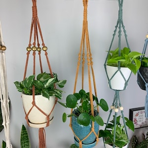 May include: Five macrame plant hangers with different colored cords and beads. The hangers are holding various plants in pots.
