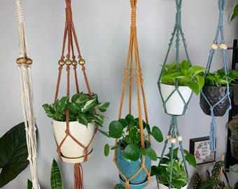 May include: Five macrame plant hangers in cream, terracotta, mustard yellow, sage green, and dusty blue, each featuring knotted cord designs, wooden beads, and various potted green houseplants suspended at different heights.