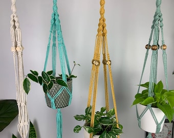 May include: Four macrame plant hangers in cream, turquoise, mustard yellow, and sage green. Each hanger features intricate knotted cord patterns, wooden beads, and holds a potted plant, except for the empty cream-colored hanger on the left.