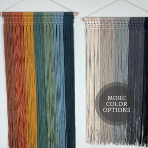 May include: Two macrame wall hangings. One features vertical stripes in shades of rust, gold, green, blue, and navy. The other has stripes in beige, gray, and black. Both are suspended from wooden dowels.