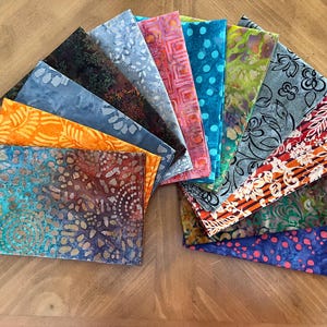 May include: A collection of rectangular fabric swatches arranged in a fan shape. The pieces feature various batik patterns including floral, geometric, and abstract designs in a vibrant palette of blue, orange, pink, teal, green, and black.