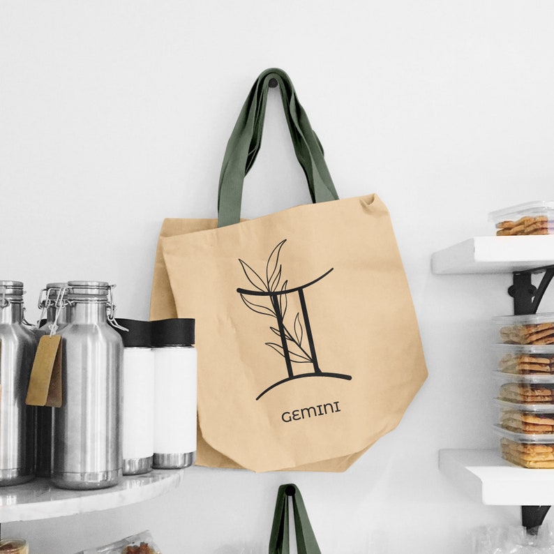 K&ouml;nnte beinhalten: Eine beigefarbene Tragetasche mit gr&uuml;nen Henkeln h&auml;ngt an einer wei&szlig;en Wand. Die Tasche zeigt ein schwarzes Zwillinge-Sternzeichen mit Bl&auml;ttern und das Wort "GEMINI" darunter. Weitere Artikel auf den Regalen sind Gl&auml;ser und Beh&auml;lter.