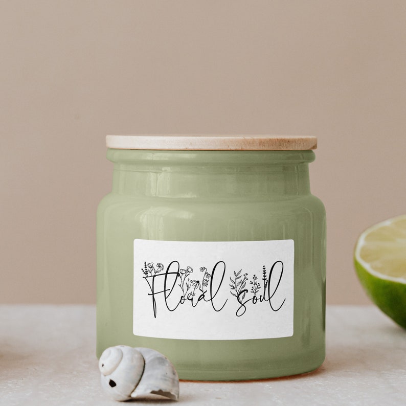 K&ouml;nnte beinhalten: Ein gr&uuml;nes Kerzengef&auml;&szlig; aus Keramik mit einem Holzdeckel und einem wei&szlig;en Etikett mit der Aufschrift "Floral Soul" und einem floralen Design.