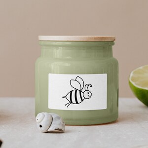 May include: A green ceramic jar with a wooden lid and a white label featuring a black and white cartoon bee. The jar is sitting on a white surface.