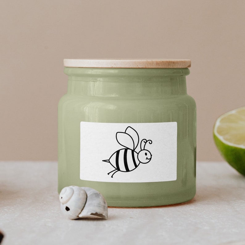 May include: A green ceramic jar with a wooden lid and a white label featuring a black and white cartoon bee. The jar is sitting on a white surface.