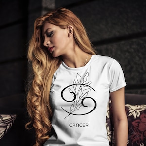 K&ouml;nnte beinhalten: Wei&szlig;es T-Shirt mit schwarzem Krebs-Sternzeichen-Symbol und floralem Design. Das Wort "CANCER" ist darunter gedruckt. Das T-Shirt hat einen Rundhalsausschnitt.