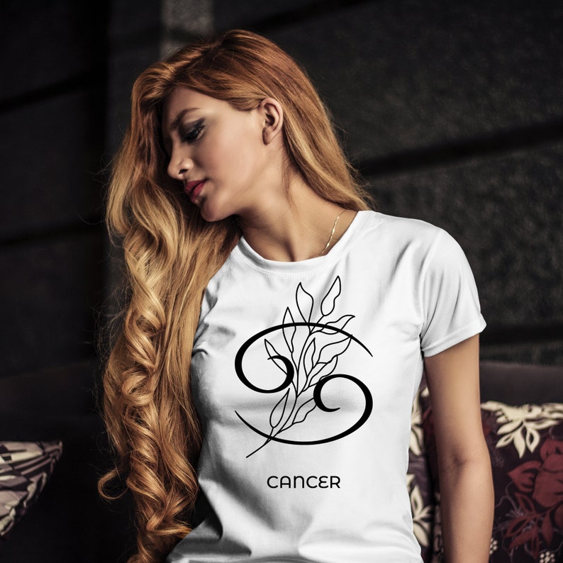 K&ouml;nnte beinhalten: Wei&szlig;es T-Shirt mit schwarzem Krebs-Sternzeichen-Symbol und floralem Design. Das Wort "CANCER" ist darunter gedruckt. Das T-Shirt hat einen Rundhalsausschnitt.