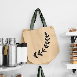 May include: A beige tote bag with a black leafy design. The bag has olive green straps and is hanging on a white wall.