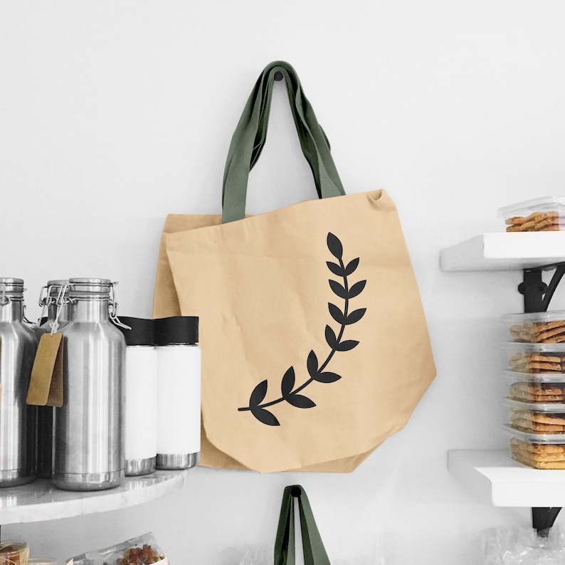 May include: A beige tote bag with a black leafy design. The bag has olive green straps and is hanging on a white wall.
