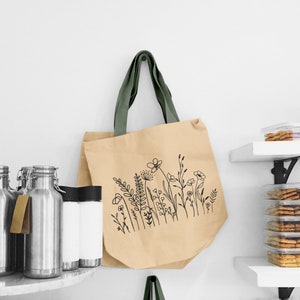 Puede incluir: Una bolsa de tela beige con un asa de tela verde y un dibujo de l&iacute;nea negra de flores silvestres en la parte delantera.