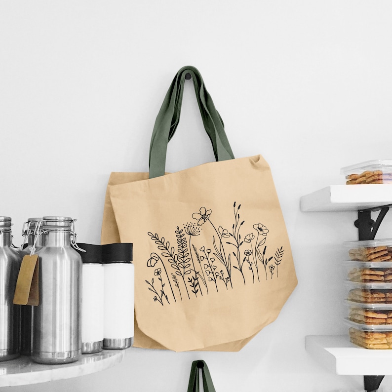 Puede incluir: Una bolsa de tela beige con un asa de tela verde y un dibujo de l&iacute;nea negra de flores silvestres en la parte delantera.