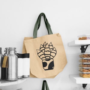 May include: A beige tote bag with green straps features a black and white illustration of a potted plant. The plant has a striped pattern and is in a black pot with a white design.