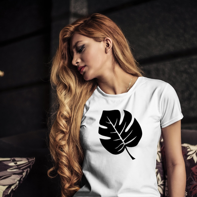 Puede incluir: Una camiseta blanca con una silueta negra de una hoja de monstera impresa en la parte delantera.