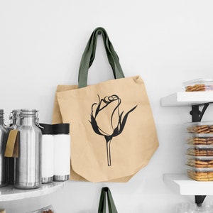 May include: A beige tote bag with green straps and a black line drawing of a rose on the front.