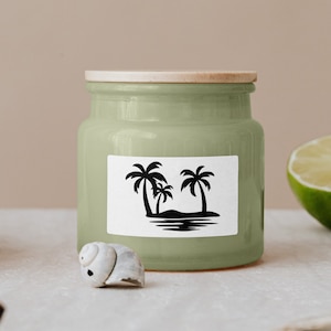 K&ouml;nnte beinhalten: Eine gr&uuml;ne Keramikdose mit einem Holzdeckel und einem wei&szlig;en Aufkleber mit einer Silhouette von Palmen auf einer Insel. Die Dose steht auf einer hellbraunen Oberfl&auml;che.