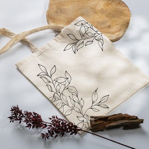 May include: A beige canvas tote bag with black line drawings of leaves and branches. The bag has a natural cotton fabric texture and a long, beige strap.
