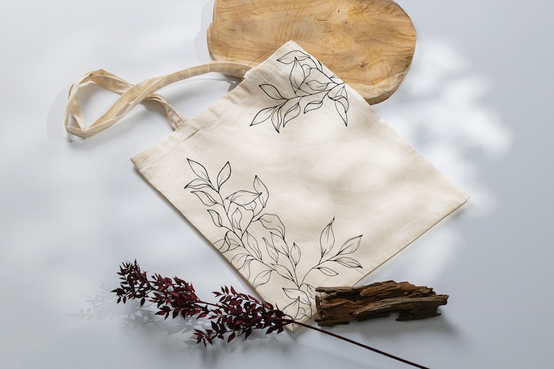 May include: A beige canvas tote bag with black line drawings of leaves and branches. The bag has a natural cotton fabric texture and a long, beige strap.