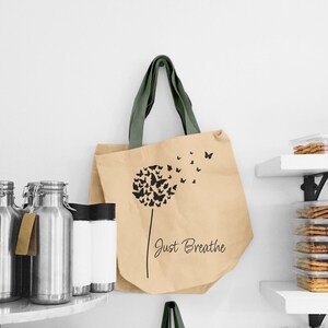 K&ouml;nnte beinhalten: Eine braune Einkaufstasche mit gr&uuml;nen Henkeln zeigt eine schwarze Silhouette eines L&ouml;wenzahns mit wegfliegenden Schmetterlingen. Der Text "Just Breathe" ist auf die Tasche gedruckt.