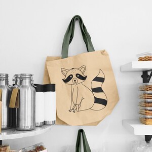 May include: A beige tote bag with a green fabric handle and a black and white illustration of a raccoon on the front. The raccoon is sitting with its tail curled around its body.