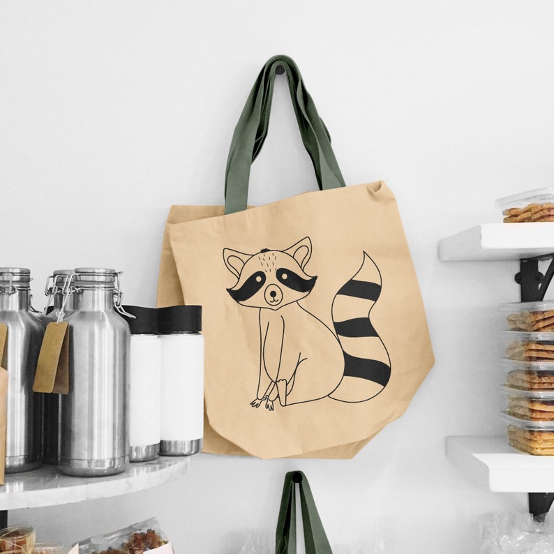 May include: A beige tote bag with a green fabric handle and a black and white illustration of a raccoon on the front. The raccoon is sitting with its tail curled around its body.