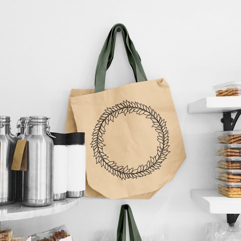 May include: A brown paper tote bag with a green fabric handle. The bag has a black wreath design with a circle in the center.