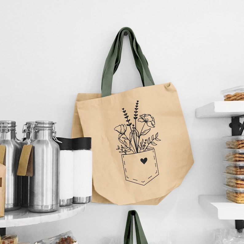 K&ouml;nnte beinhalten: Eine beige Tragetasche mit gr&uuml;nen Henkeln und einer schwarzen Linienzeichnung von Blumen in einer Tasche mit einem Herzen. Die Tasche ist aus einem nat&uuml;rlichen Material gefertigt und hat ein schlichtes, minimalistisches Design.