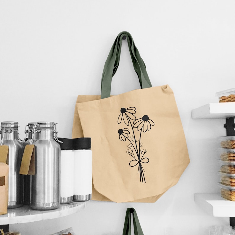 May include: A beige canvas tote bag with a black floral design of daisies and green straps. The bag is hanging on a wall. Several stainless steel bottles and jars are on a shelf in the background.