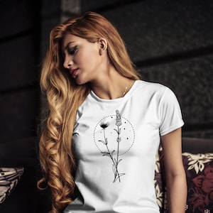 May include: White t-shirt featuring a black floral design with a circle of stars. The design includes a dandelion and lavender sprig. The shirt has short sleeves and a crew neck.