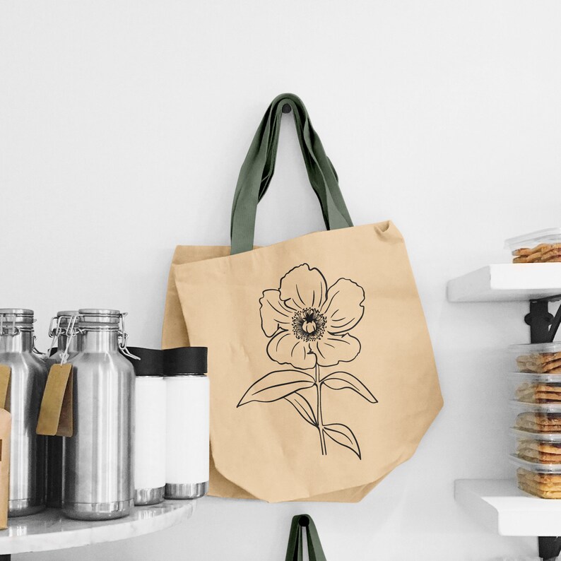 Puede incluir: Una bolsa de papel marr&oacute;n con asas verdes presenta un dibujo de l&iacute;nea negra de una flor de cinco p&eacute;talos. La bolsa est&aacute; colgada en una pared blanca.