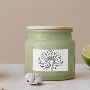 Puede incluir: Un tarro de vela de cer&aacute;mica verde con tapa de madera y una etiqueta blanca con un dibujo de l&iacute;nea negra de una margarita.