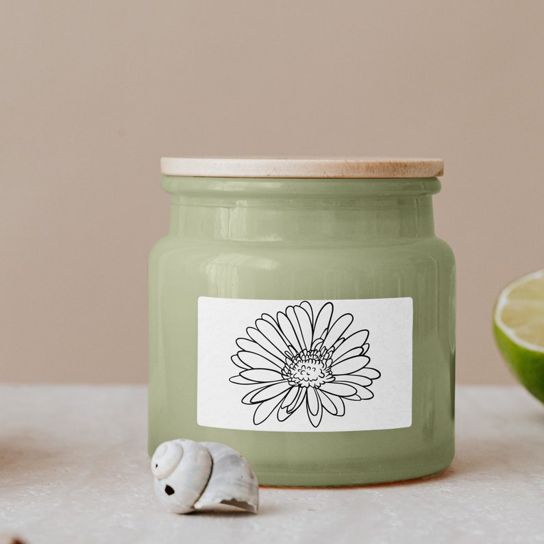 Puede incluir: Un tarro de vela de cer&aacute;mica verde con tapa de madera y una etiqueta blanca con un dibujo de l&iacute;nea negra de una margarita.