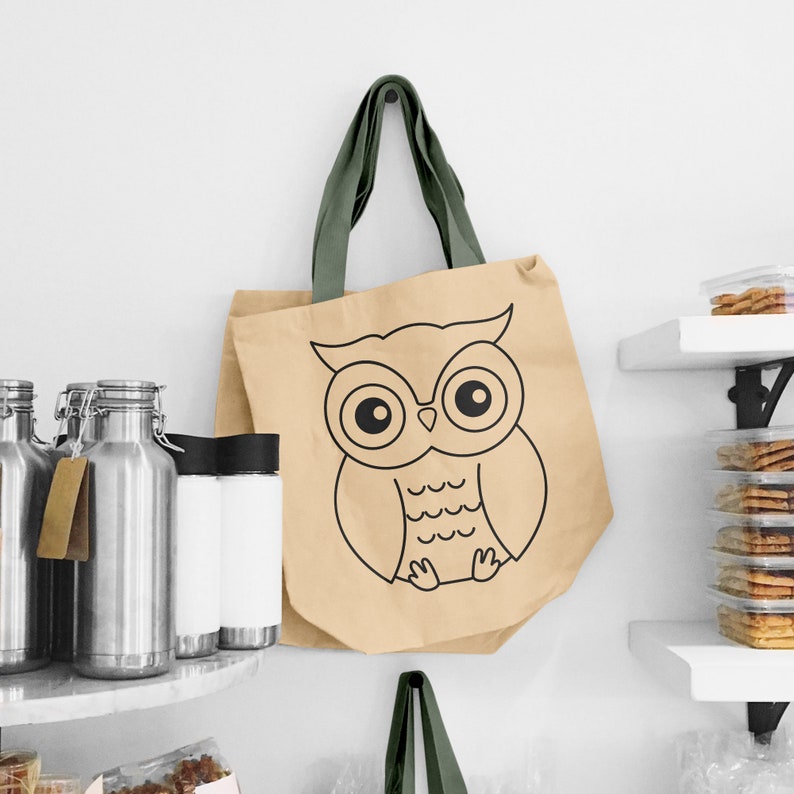 May include: A beige tote bag with green handles features a black and white outline of an owl with large eyes. The owl is sitting on a branch with its wings spread out.