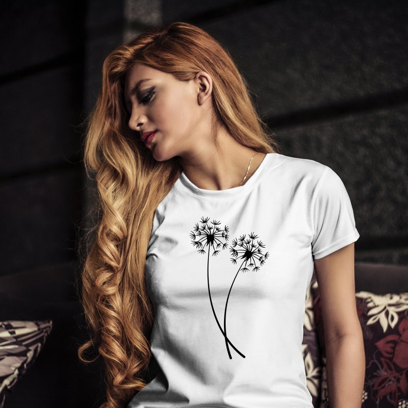 K&ouml;nnte beinhalten: Ein wei&szlig;es T-Shirt mit einem schwarzen L&ouml;wenzahnmuster. Das L&ouml;wenzahnmuster zeigt zwei L&ouml;wenz&auml;hne, deren Samen im Wind fliegen.
