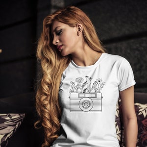 K&ouml;nnte beinhalten: Wei&szlig;es T-Shirt mit einer schwarzen Linienzeichnung einer Kamera, aus der Blumen wachsen.