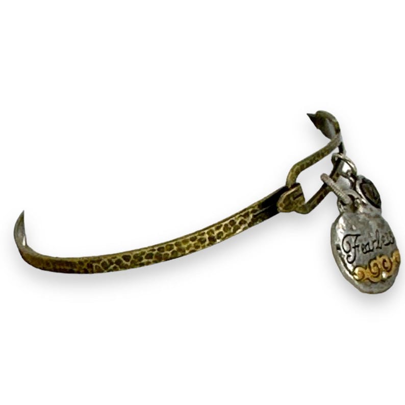 May include: A hammered brass-tone bangle bracelet with a silver-tone charm. The charm is heart-shaped with the word "Fearless" in script and gold accents. The bracelet has a hook closure.