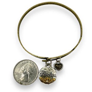 May include: A brass-toned bangle bracelet with two charms and a quarter coin for size comparison. One charm is a textured silver disc with the word "Fearless" and gold accents. The other is a heart charm with the initials "B" and "B".