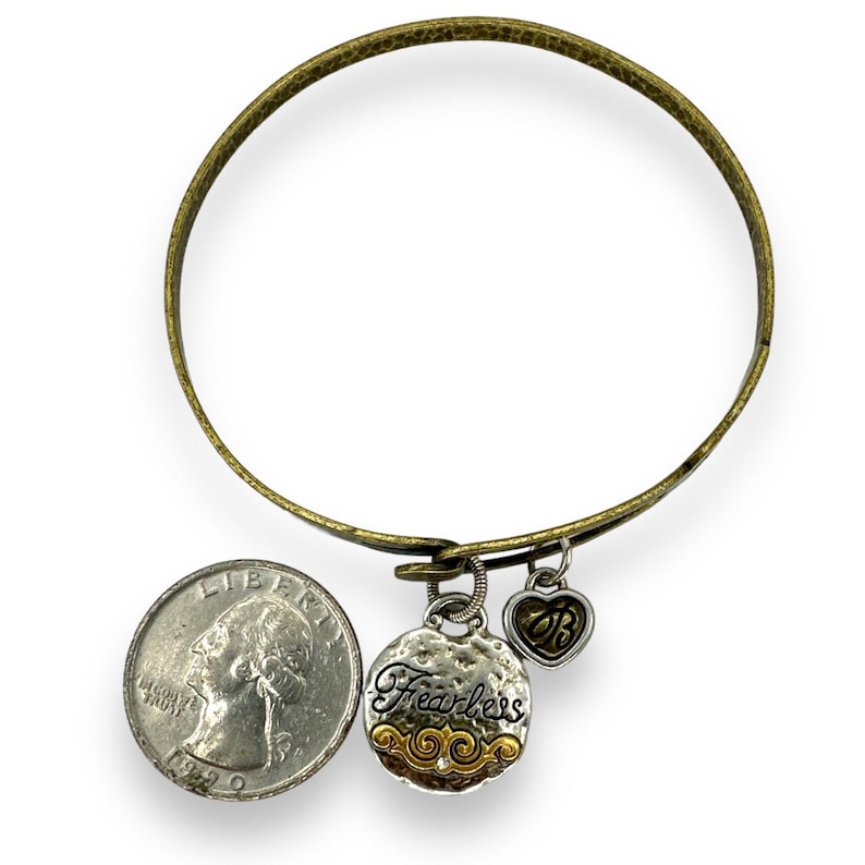 May include: A brass-toned bangle bracelet with two charms and a quarter coin for size comparison. One charm is a textured silver disc with the word "Fearless" and gold accents. The other is a heart charm with the initials "B" and "B".