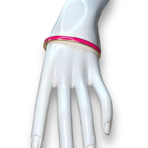 Puede incluir: Un brazalete dorado con una franja de esmalte rosa brillante. El brazalete se exhibe en una mano de maniquí blanca, mostrando su diseño y contraste de color. La pieza de joyería es un accesorio simple y elegante.