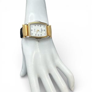 May include: A rectangular gold-toned watch with a white face and black numerals. The watch has a black leather band and is displayed on a white mannequin hand. The watch face displays the numbers 1 through 12, with gold-toned hands.