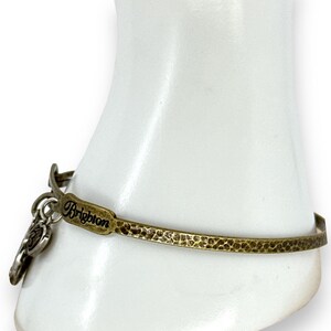 May include: A gold-toned metal bracelet with a hammered texture. The bracelet features the word "Brighton" and a charm. The bracelet is displayed on a white form.