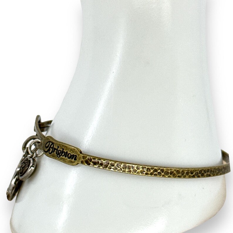 May include: A gold-toned metal bracelet with a hammered texture. The bracelet features the word "Brighton" and a charm. The bracelet is displayed on a white form.