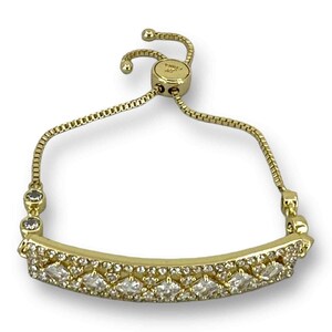 May include: Gold-toned bracelet with a row of clear, sparkling stones. The bracelet features a delicate chain and a sliding clasp for adjustable sizing. The main design element is a curved bar adorned with multiple clear, faceted stones.