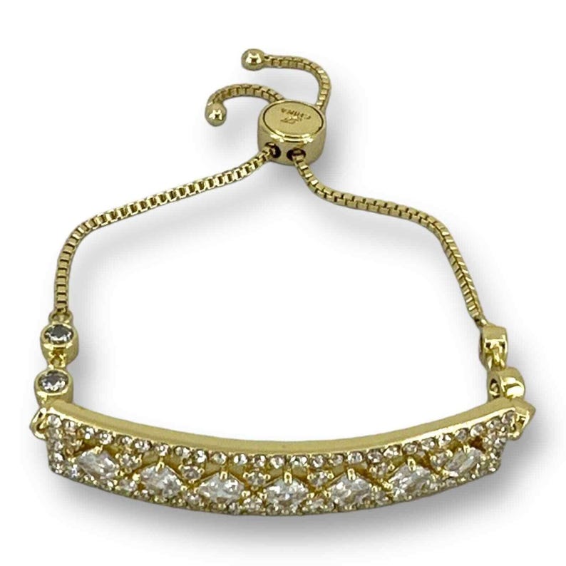 May include: Gold-toned bracelet with a row of clear, sparkling stones. The bracelet features a delicate chain and a sliding clasp for adjustable sizing. The main design element is a curved bar adorned with multiple clear, faceted stones.