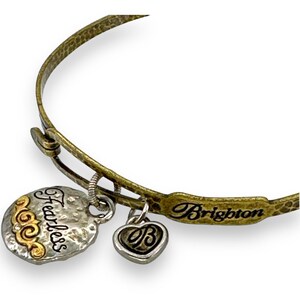 May include: A gold-toned metal bangle bracelet with three charms. One charm is a silver-toned heart with a 'B' in the center. Another is a silver-toned disc with the word 'Fearless' and gold accents. The bracelet is marked 'Brighton'.