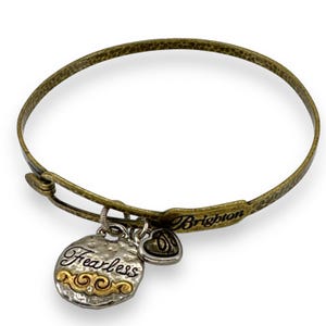 May include: A gold-toned metal bangle bracelet with a textured finish. The bracelet features a charm with the word "Fearless" and a heart-shaped charm. The brand name "Brighton" is visible on the bracelet.