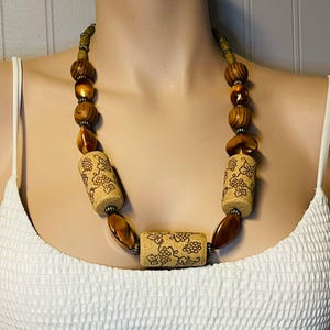 Op de afbeelding: Een ketting met houten kralen in bruine en amberkleurige tinten. Het heeft kurkkralen met een druivenrankpatroon. De ketting wordt gepresenteerd tegen een witte achtergrond. De ketting is ongeveer 45 cm lang.