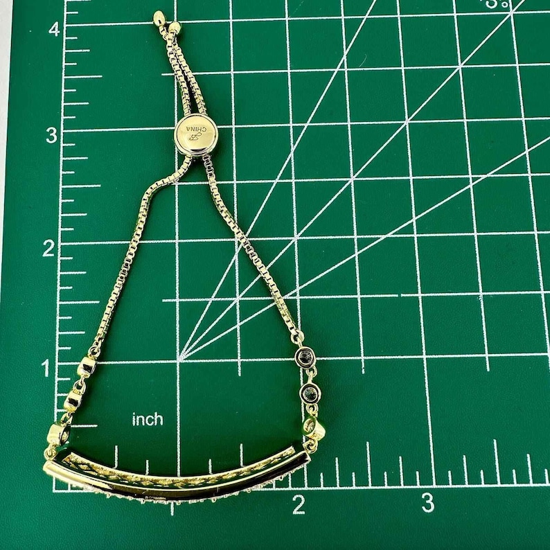May include: Gold-toned adjustable bracelet with a curved bar design. The bar is embellished with small, clear crystals. The bracelet has a sliding bead for adjustment and a small charm that says "CHINA". The bracelet measures approximately 2 inches in length.