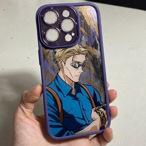 Pu&ograve; includere: Una custodia per telefono viola con un designo astratto oro e nero. La custodia presenta un'immagine stampata di un uomo con i capelli biondi che indossa occhiali e una camicia blu.