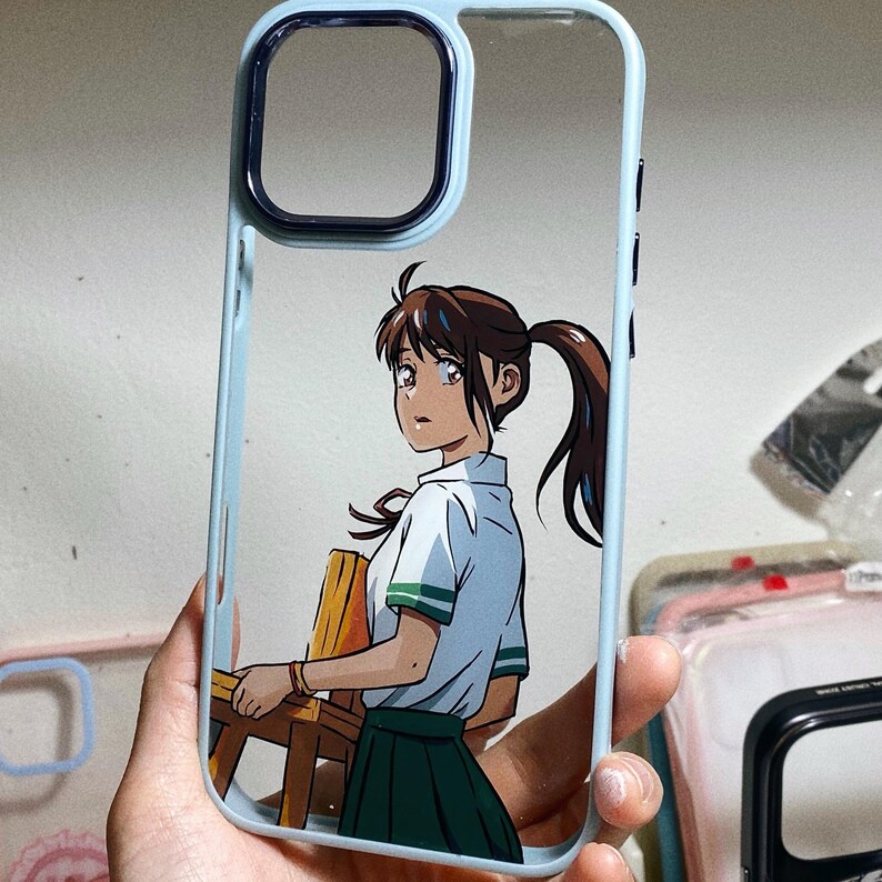Pu&ograve; includere: Una custodia per telefono trasparente con un bordo blu chiaro e un'immagine stampata di un personaggio anime con capelli castani e un'uniforme scolastica blu e verde. Il personaggio tiene una sedia di legno.