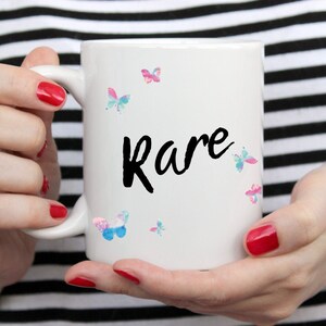 May include: White ceramic mug with the word "Rare" in black script, decorated with watercolor butterfly illustrations in shades of pink, blue, and teal. The mug is held by a person with red nail polish.